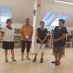 Štiri ženske stojijo v vrsti v prostoru z knjižno polico in mizami v ozadju. Ena izmed njih drži v roki zvezek. Vse zrejo naprej, kot da nastopajo ali govorijo občinstvu.