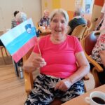 Starejša ženska v roza majici, ki veselo drži ročno izdelano slovensko zastavo, sedi za mizo; pred njo je skodelica kave.