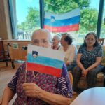 Starejša ženska drži ročno narisano slovensko zastavo, v ozadju več drugih stanovalcev in velika slovenska zastava ob oknu.
