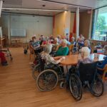 Skupina starejših oseb na invalidskih vozičkih in stolih sedi zbrana okoli miz, poslušajo nastop ali govor ob praznovanju državnega praznika. V ozadju je velika slovenska zastava.