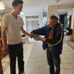 Zaposleni moški v beli majici izroča časopis stanovalcu doma, ki nosi modro jakno. Nahajata se v notranjosti doma starejših.
