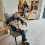 Starejši moški sedi v naslanjaču in bere časopis. V ozadju je računalniški kotiček, kjer tri osebe delajo za mizo.