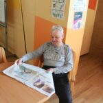 Starejši moški sedi za leseno mizo in bere časopis. V ozadju so oranžne stene z informacijskimi plakati.