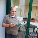 Moški srednjih let stoji pred vhodom v dom in bere revijo Radovednež, v ozadju steklena vrata in cvetlična okenska polica.