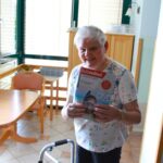 Starejša gospa nasmejano stoji z revijo Radovednež v rokah in jo ponosno kaže proti kameri v notranjosti doma.