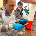 Moški z modrimi zaščitnimi rokavicami in plastičnim predpasnikom slika kartonski valj z rdečo barvo, medtem ko drugi uporabniki doma za starejše opazujejo.
