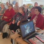 Skupina starejših oseb, ki sedi v krogu in gleda film na prenosnem računalniku; na zaslonu je prizor iz filma Kekec.