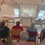 Skupina stanovalcev doma za starejše, večinoma s sivimi lasmi, sedi na stolih in v invalidskih vozičkih ter gleda slovenski film Kekec, ki se predvaja na velikem projekcijskem platnu.