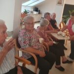 Stanovalci doma za starejše med ogledom filma Kekec sedijo na stolih in pijejo iz papirnatih lončkov, medtem ko zaposlena v rdeče-beli uniformi deli napitke.
