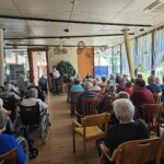 Pogled s hrbta na skupino stanovalcev doma za starejše, ki poslušajo nastopajočega moškega na odru v večnamenskem prostoru.