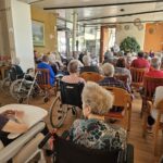 Starejši stanovalci, večinoma v invalidskih vozičkih, sedijo v dvorani in spremljajo kulturni program z nastopajočim moškim na odru.