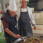 Dva nasmejana kuharja pozirata pred veliko ponevjo, polno barvite zelenjavne jedi. Eden drži lopatko, drugi mu z roko prijateljsko počiva na rami.