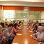 Skupina starejših žensk sedi ob dolgi mizi in uživa sadne sladice. V ozadju je svetla jedilnica z velikimi okni in oranžnimi zavesami.