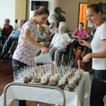 Dve zaposleni pripravljata ledeno kavo za skupino starejših oseb, ki sedijo v ozadju na invalidskih vozičkih in čakajo na postrežbo.
