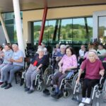 Skupina stanovalcev v invalidskih vozičkih sedi v senci pred domom in spremlja dogajanje.