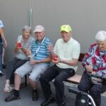 Pet stanovalcev doma sedi ob steni zunaj, v rokah držijo kozarce z limonado in se sproščeno družijo.