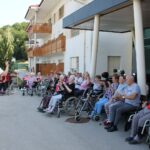 Stanovalci doma upokojencev sedijo zunaj pred vhodom in izvajajo vodeno vadbo z dvignjenimi rokami.