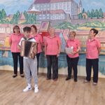 Pet žensk v roza srajcah in deček z diatonično harmoniko nastopajo pred poslikano steno z motivom mesta.