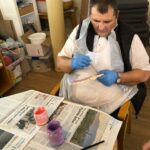 Starejši moški sedi za mizo, oblečen v zaščitni predpasnik in modre rokavice, medtem ko natančno barva leseno ribo z vijolično in rožnato barvo.