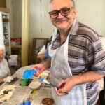 Nasmejan starejši moški z očali drži čopič in pobarvano leseno ribo v roki, obkrožen z barvnimi kozarčki in ostalimi udeleženci delavnice.