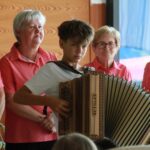 Mlajši deček igra diatonično harmoniko pred skupino starejših žensk, ki so oblečene v usklajene rožnate srajce.