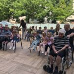 Otroci in starejši sedijo skupaj v krogu na terasi. Medgeneracijsko druženje poteka v prijetnem ozračju ob drevesih.