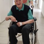 Nasmejan starejši gospod sedi v invalidskem vozičku, za njim stoji mlada deklica, ki ga potiska po hodniku doma za starejše.