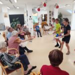 Skupina starejših oseb sedi na stolih v polkrogu in meče pisane žogice v pladnje, ki jih držijo otroci, v svetlem prostoru doma za starejše.