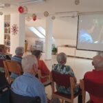Starejši gledajo film na platnu v večnamenskem prostoru, na zaslonu je prizor z nasmejano deklico.
