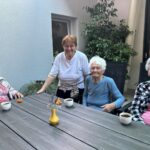 Štiri starejše ženske sedijo za vrtno mizo v senci in se družijo. Na mizi so skodelice, koščki sadja in okrasna vaza z rožami.