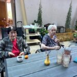 Dve starejši ženski sedita na dvorišču doma starejših. Na mizi pred njima so kozarci, vrč s pijačo, skodelici s kavo in koščki sadja.