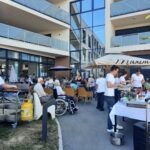 Veliko število stanovalcev doma za starejše sedi na terasi sodobne stavbe. Kuharsko osebje pripravlja hrano na žaru in streže ob mizah pod senčniki.