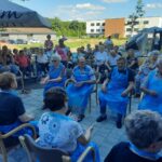 Starejši stanovalci, oblečeni v modre zaščitne predpasnike, sodelujejo v skupinski aktivnosti na prostem, obkroženi z drugimi opazovalci in osebjem.