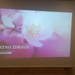 Na fotografiji je projekcija predstavitve z naslovnim diapozitivom »USTNO ZDRAVJE«. V ozadju je mehka, rožnato vijolična fotografija belega cveta, ki daje pomirjujoč ton predstavitvi. Avtorica je diplomirana medicinska sestra Katja Golob, v spodnjem levem kotu pa je logotip Zdravstvenega doma Koper. Desno spodaj je letnica dogodka: Koper 2025. Fotografija prikazuje uvod v izobraževanje o ustni higieni, namenjeno predvsem starejšim.