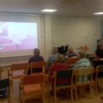 Fotografija prikazuje predavateljico, ki stoji levo od projekcije, kjer je prikazan začetni diapozitiv z naslovom »USTNO ZDRAVJE«. V dvorani sedi več kot deset udeležencev, pretežno starejših, ki spremljajo predstavitev. Stoli so razporejeni v vrstah, več oseb uporablja bergle ali invalidske vozičke. Prostor je osvetljen z umetno svetlobo, vzdušje pa je umirjeno in osredotočeno. Predavateljica je oblečena v svetlo modro majico in temne hlače.