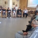 Skupina stanovalcev doma za starejše sedi v krogu v skupnem prostoru in sodeluje v pogovoru ali delavnici.