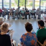 Velika skupina stanovalcev doma za starejše sedi v krogu in sodeluje pri skupinski dejavnosti ali druženju.