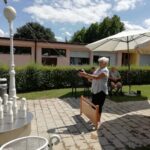 Starejša gospa na prostem izvaja met ruske kegljaške krogle proti kegljem na posebnem stojalu; v ozadju sedijo stanovalci doma za starejše pod senčniki.