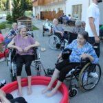 Dve starejši ženski v invalidskih vozičkih namakata noge v plastični bazenček, v ozadju več drugih stanovalcev in zaposlenih pred stavbo doma starejših.