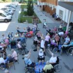 Večja skupina stanovalcev doma za starejše osebe se udeležuje aktivnosti na prostem, sedečih v invalidskih vozičkih in stolih, z dvignjenimi rokami, obkroženi z osebjem in plastičnimi bazenčki z vodo.