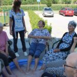 Skupina starejših žensk ob robu parkirišča sedi okoli napihljivega bazenčka in namaka noge v vodi, ob njih stoji zaposlena.