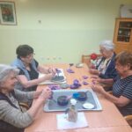 Štiri starejše ženske sede za mizo ročno izdelujejo vijoličaste sivkine srčke iz blaga.