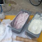 Pogled na voziček s postreženim sladoledom – dva velika kovinska zabojnika z vaniljevim in jagodnim sladoledom, prazni korneti in papirnate skodelice ob strani. V ozadju vidna kolesa invalidskega vozička.