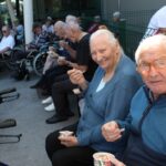 Starejša ženska in moški sedita zunaj in jesta sladoled iz lončka, ženska se veselo smeje proti kameri.