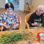 Starejša ženska in moški sedita za mizo in ročno trgata liste mete za pripravo čaja v notranjih prostorih doma.