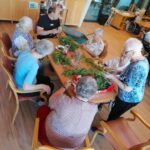 Več stanovalcev doma za starejše, večinoma žensk, sedi okoli mize in obdeluje meto; na mizi je veliko svežih zelišč in kozarec z napitkom.