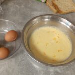 Na kuhinjskem pultu so skleda z razžvrkljanimi jajci in mlekom, cela jajca v prozorni posodi, jajčne lupine na papirju ter rezine kruha pripravljene za namakanje.