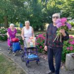 Trije stanovalci doma za starejše sprehajajo med hortenzijami, eden izmed njih v ospredju drži šopek cvetja.