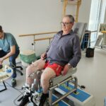 Starejši moški izvaja vadbo za noge na rehabilitacijski napravi, ob njem sedi terapevt z opremo.