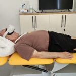 Starejši moški leži na terapevtski mizi z VR očali na glavi, uporablja virtualno resničnost za terapijo.